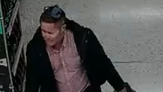 A grainy CCTV still image showing a middle-aged man in a pink button up shirt with a collar and a dark jacket. He is pulling along a bag of shopping in a supermarket.