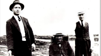 A black and white picture of three men in a field wearing hats and suits. Two younger men are standing either side of an older man who is sitting down.