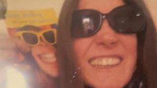 A woman wearing dark sunglasses smiles at the camera. She is holding up a picture of a monkey smiling and wearing sunglasses next to her face. 