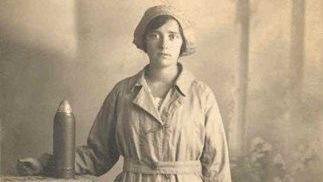 Black and white photograph of a woman, thought to be that of Gloucester Canary Girl Nell Slatter. She is wearing the working overalls and hat of the National Filling Factory No. 5 which was established at Quedgeley. There is a large shell cartridge sitting on a table next to her.