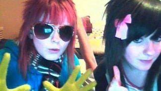 A girl with short pink hair and a straight fringe wears large aviator sunglasses. She has yellow gloves on and is wearing a bright blue hoodie and a scarf. To the right, a girl with black hair with a pink bow gives a thumbs up. 