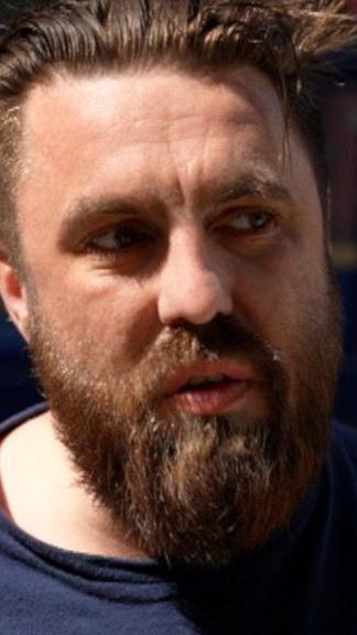 A man with brown hair and a brown beard is wearing a blue t-shirt.