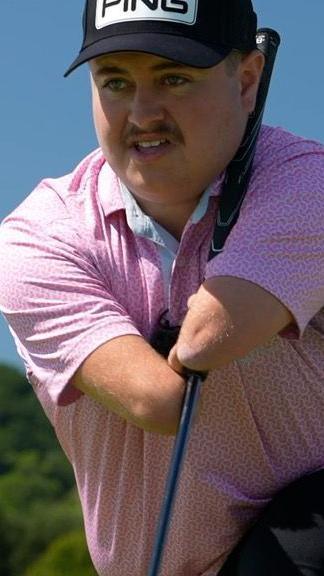 A young golfer with no hands kneels to assess the golf course, wearing a pink polo shirt and black baseball cap.