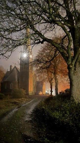 A tall church with a lit clock tower stands at the end of a dim, tree-lined path.