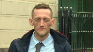 Craig Rowland - a still of a man with short brown hair wearing a light blue collared shirt, a skinny grey tie and a dark jacket which is open. He is walking in front of a tall, black gate and a portland stone wall is in the background