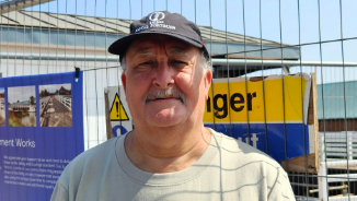 An older man wearing a dark cap with a logo on and a light grey top stands in front of safety barriers at a canal bridge, which is closed for repair works. He has short grey hair and a moustache. There are signs warning people of danger and updating them on the works behind him.