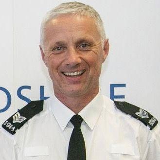 Dave Cruxton, a man wearing a white police shirt with black police epaulettes bearing his force ID number. He is smiling, posing for the camera.