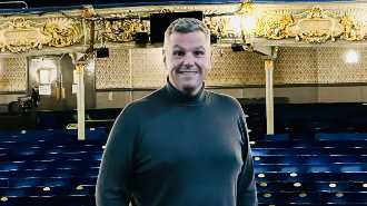 Jonathan Higgins, with short grey hair, wearing a grey long-sleeve top and black pants and trainers, has his hands in his pockets as he stands on a stage. There are seats behind him.