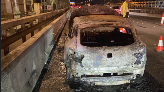 A burnt car parked on a motorway carriageway with its bonnet lifted, and a traffic officer standing beside it.