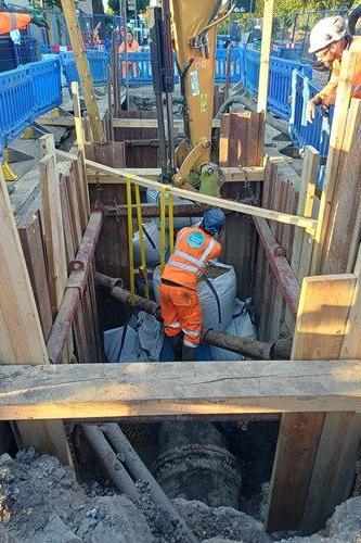 Engineers carrying out repair work in a deep excavated trench around a large water main. A worker in high-visibility orange protective clothing stands at the bottom of the shored excavation beside exposed pipework and repair materials, while another worker operates machinery from the surface.