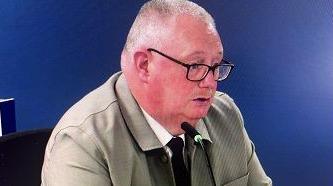 Greogory McQuitty has short grey hair and is wearing black-rimmed glasses. He is wearing a grey jacket, white shirt and black tie.