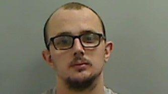 Mugshot of Cooper. He has a blading head of dark hair, dark goatee and moustache with black glasses