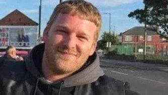 Mr Sutton smiles at the camera, wearing what appears to be a leather vest with a dark hoodie underneath. He has strawberry blond hair and stubble on his face. A red brick house and the road can be seen on the right in the background and a boohoo billboard on the left.