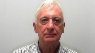 Man with white hair wearing a checked shirt looks into a camera.