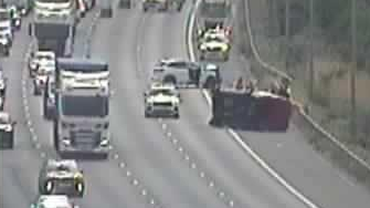 A still taken from a grainy traffic camera. It shows a dark van on its side with people around it. Police cars have blocked it in and traffic is passing to the left.