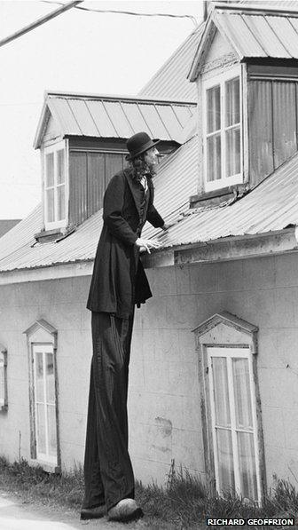 Gilles Ste Croix in stilts in front of house