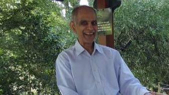 Bhim Kohli smiling and wearing a light blue collared shirt. He is sat in front of green foliage