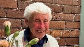 An elderly man smiles at the camera. He is wearing a white cardigan and blue shirt. He has white hair. He is sitting against a brick wall, flowers obscure his face.