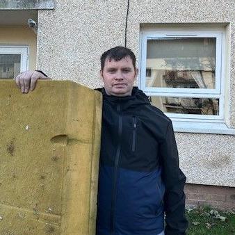 Andrew Bond, wearing a dark jacket with a lighter grey panel, stands with the mattress he jumped on to from his balcony to escape a fire in Niddrie Mill. The mattress is yellow sponge and has some staining. The flat block is behind him.