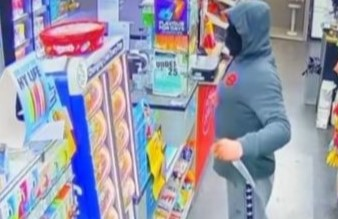 A man dressed in a tracksuit holding a kitchen knife. He has concealed his face and is staring across the counter.