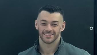 Josh Irwin is standing behind a black backdrop and is smiling. 
He has short black hair and is wearing a grey jacket and black T-shirt. 