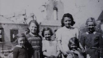 A group of nine children, with their faces obscured by gray rectangles, are standing and sitting in front of a backdrop of partially destroyed buildings. Some children are crouching or sitting on the ground, while others are standing. The scene suggests a setting affected by war or conflict.