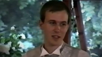 Leon Adams before the attack. He has close cropped brown hair and is wearing a grey suit, a white collar is visible. The background is white roses and other greenery. 