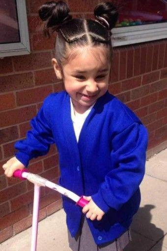 Shay as a younger child in a blue school uniform on a pink scooter, grinning