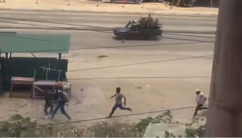 Men running along a road chased by an open backed vehicle with armed police