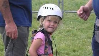 Elana is in the middle looking towards the camera smiling. She has blonde hair and a fringe and is wearing a white helmet. She has a pink top on with a harness over the top.