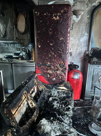 A charred, melted red fridge and a burnt our sofa in front of it. There are kitchen units that are blackened and charred. 