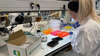 A scientist wearing a white lab coat uses a pipette to drip liquid into small viles in a bright orange tray.