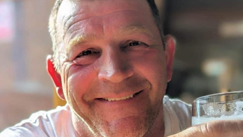 Paul Quinn, who has short light brown hair, smiles at the camera while holding a pint of what appears to be lager and wearing a white vest-top. 