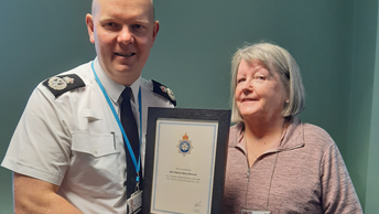 A woman and a man smile for the camera and are both holding a police certificate noting the retirement of an officer after 55 years in North Yorkshire Police. The man is wearing police attire and the woman is dressed casually.
