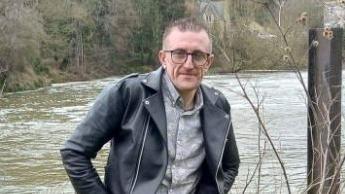 A man with short grey hair and glasses wearing a black leather jacket and grey shirt leaning on railings in front of a river