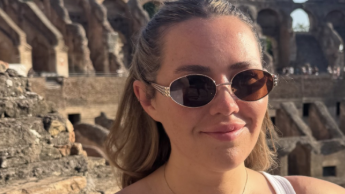 Ella Nickson is wearing sunglasses and her hair is half-tied up. She is standing inside the Colosseum in Rome