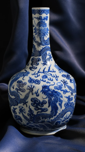 A blue and white porcelain vase, with a very slender neck and a pattern of dragons and flower-like shapes. It is set against a backdrop of a blue shiny piece of material.