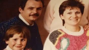 An old family posed photograph featuring a small boy about six, a woman in a cream, pink, yellow, green and brown jumper with short brown hair and a man in a dark jumper with dark hair and moustache.