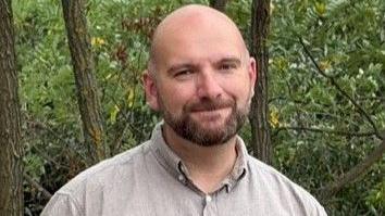 A man with a brown shirt on standing with trees and bushes behind him.