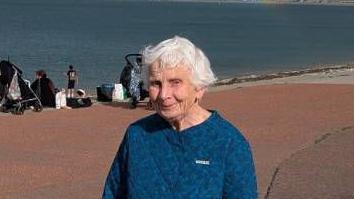 A woman with white hair, wearing a blue top and a blue and white flowery skirt is seen standing on a beach
