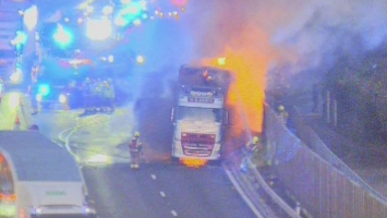 Huge orange flames are coming out of a lorry which is stopped on the shoulder of a motorway. Firefighters standing nearby are spraying it with fire hoses.