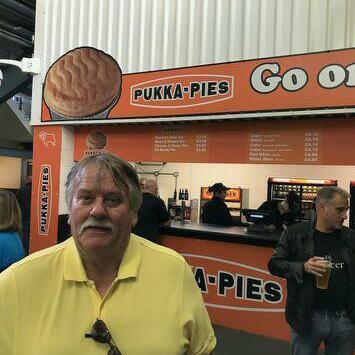 A man with grey hair and a grey moustache, wearing a yellow t-shirt, stands in front of a Pukka Pies stand with orange signs 