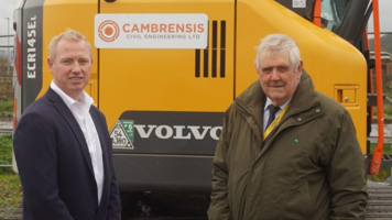 A man in a navy blue suit and white shirt stands beside a man in a dark green coat. In the background is a yellow Volvo digger and metal fences. 