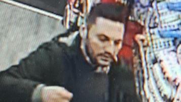 A CCTV image of a man in a shop. He has dark hair and a beard, and is wearing a dark coloured coat. 