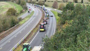 A traffic camera image of a dual carriageway. On the right hand carriageway, tractors in a convoy move toward the camera in one of the two lanes. Other vehicles travel past them in the other lane. 