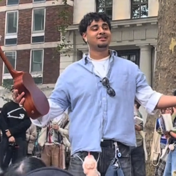 A young man holds up a ukulele after performing to a crowd in Soho Square. He is wearing a blue checked shirt, has sunglasses hanging from his top button as well as a Labubu and cap from his trousers.