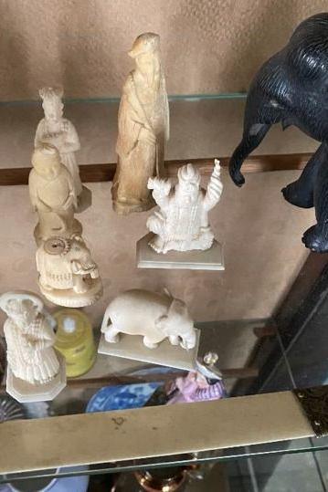 Seven white ivory ornaments including an elephant and sculpted figures and a black elephant ornament are on display on a glass shelf.
