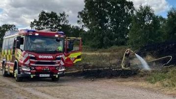 A fire engine is parked on the road next to a scorched area of heath, as a firefighter sprays water on it. There are trees in the background.