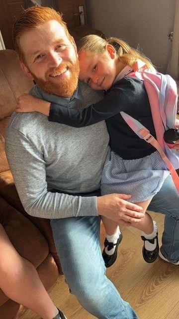 Dad Danny Jones, smiling sat down wearing jeans and a jumper, has a ginger hair and beard. His daughter sits on his knee in a school uniform, backpack and blonde pigtails. 