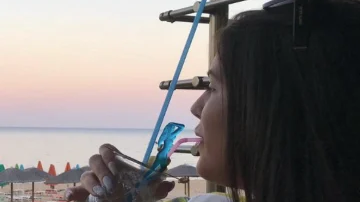 A young woman with long dark brown hair is drinking through a straw in a glass while she appears to be looking out towards the sea in a holiday resort.